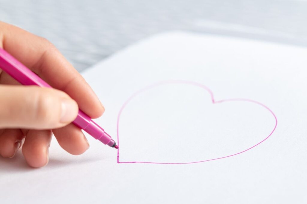 Hand zeichnet mit rosa Stift ein Herz auf weißes Papier.