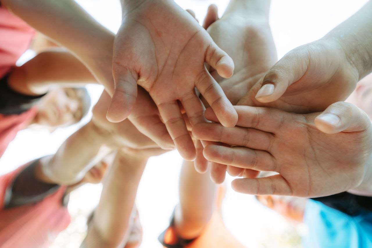 Nahaufnahme von mehreren Händen verschiedener Menschen, die in der Mitte zusammengelegt werden, vor hellem Hintergrund – ein Zeichen für Gemeinschaft und Zusammenarbeit.